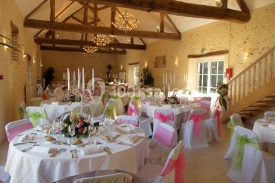 Salle de réception lumineuse avec tables rondes décorées, chaises blanches et poutres apparentes en bois.
