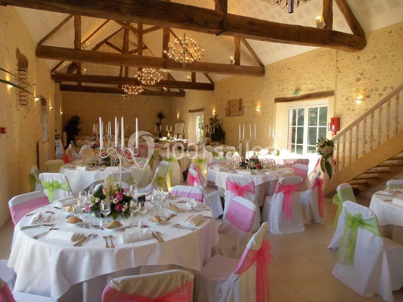 Salle de réception décorée avec des tables rondes, nappes blanches, chaises ornées de rubans colorés et chandeliers.