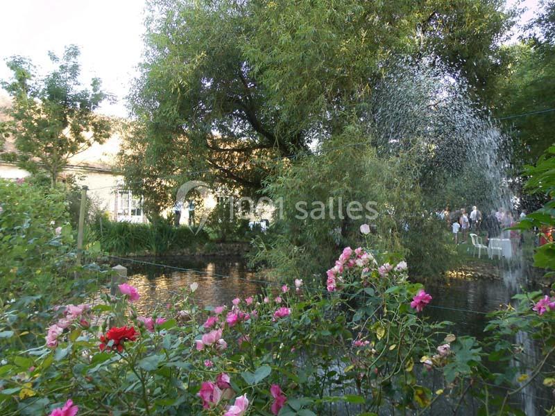 Jardin fleuri avec des rosiers, un étang, des arbres et une maison en arrière-plan sous un ciel dégagé.