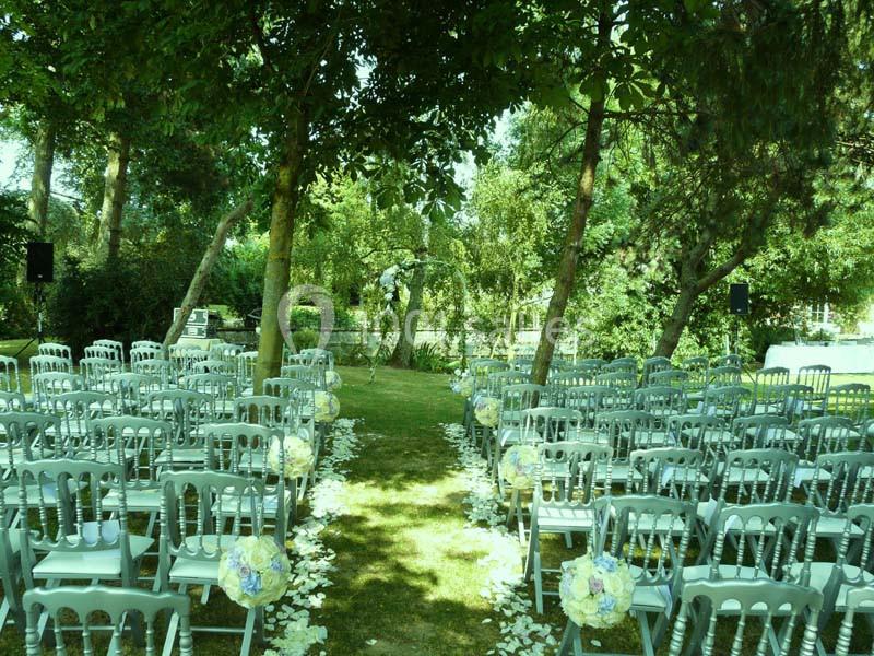Chaises alignées en extérieur sous des arbres, formant une allée centrale décorée de pétales et de bouquets pour une…