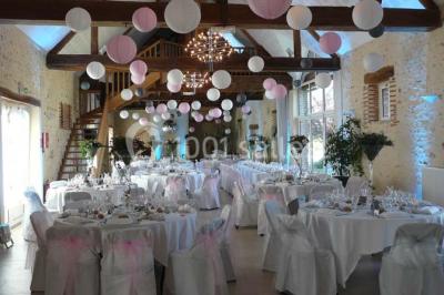 Salle de réception lumineuse avec tables rondes décorées, chaises blanches et poutres apparentes en bois.