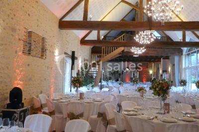 Salle de réception lumineuse avec tables rondes décorées, chaises blanches et poutres apparentes en bois.