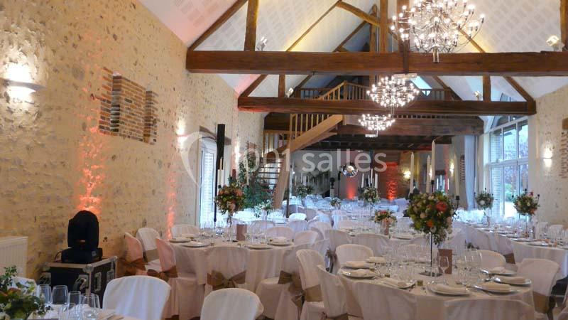 Salle de réception décorée avec des tables dressées, des chaises blanches, des chandeliers et des poutres apparentes.