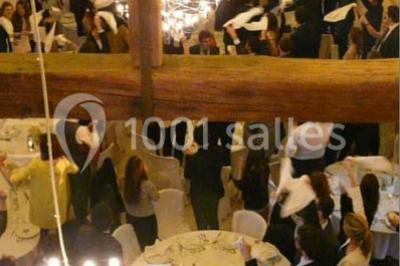 Salle de réception lumineuse avec tables rondes décorées, chaises blanches et poutres apparentes en bois.