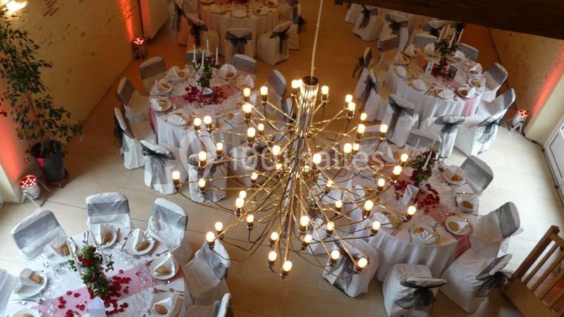 Salle de réception décorée avec des tables rondes dressées, nappes blanches, chaises ornées et lustre central lumineux.