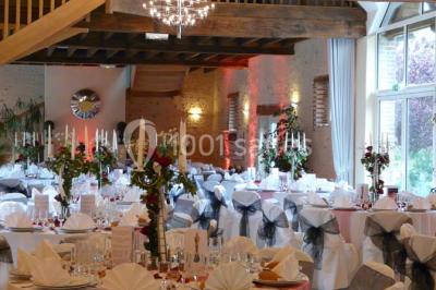 Salle de réception lumineuse avec tables rondes décorées, chaises blanches et poutres apparentes en bois.