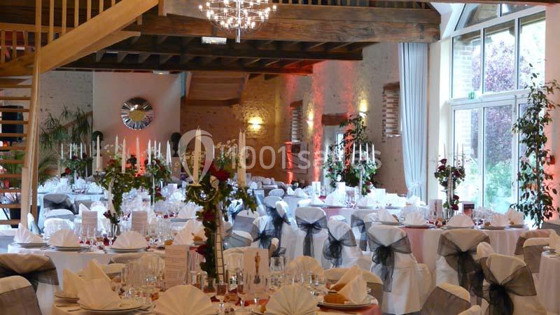 Salle de réception élégamment décorée avec des tables rondes, nappes blanches, chaises ornées de rubans noirs et chandeliers.