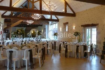 Salle de réception lumineuse avec tables rondes décorées, chaises blanches et poutres apparentes en bois.