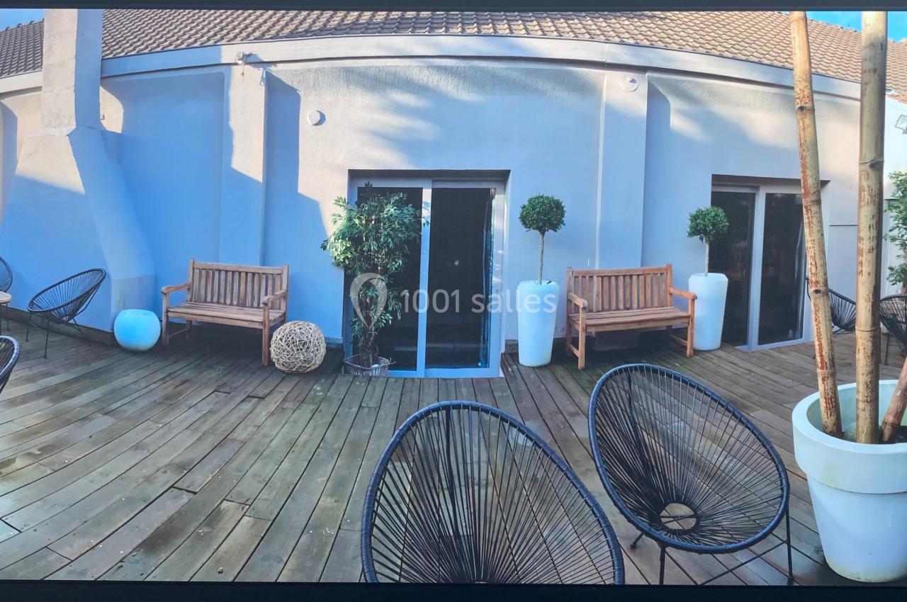 Terrasse en bois avec mobilier d'extérieur, chaises noires, bancs en bois, plantes en pots et murs blancs.