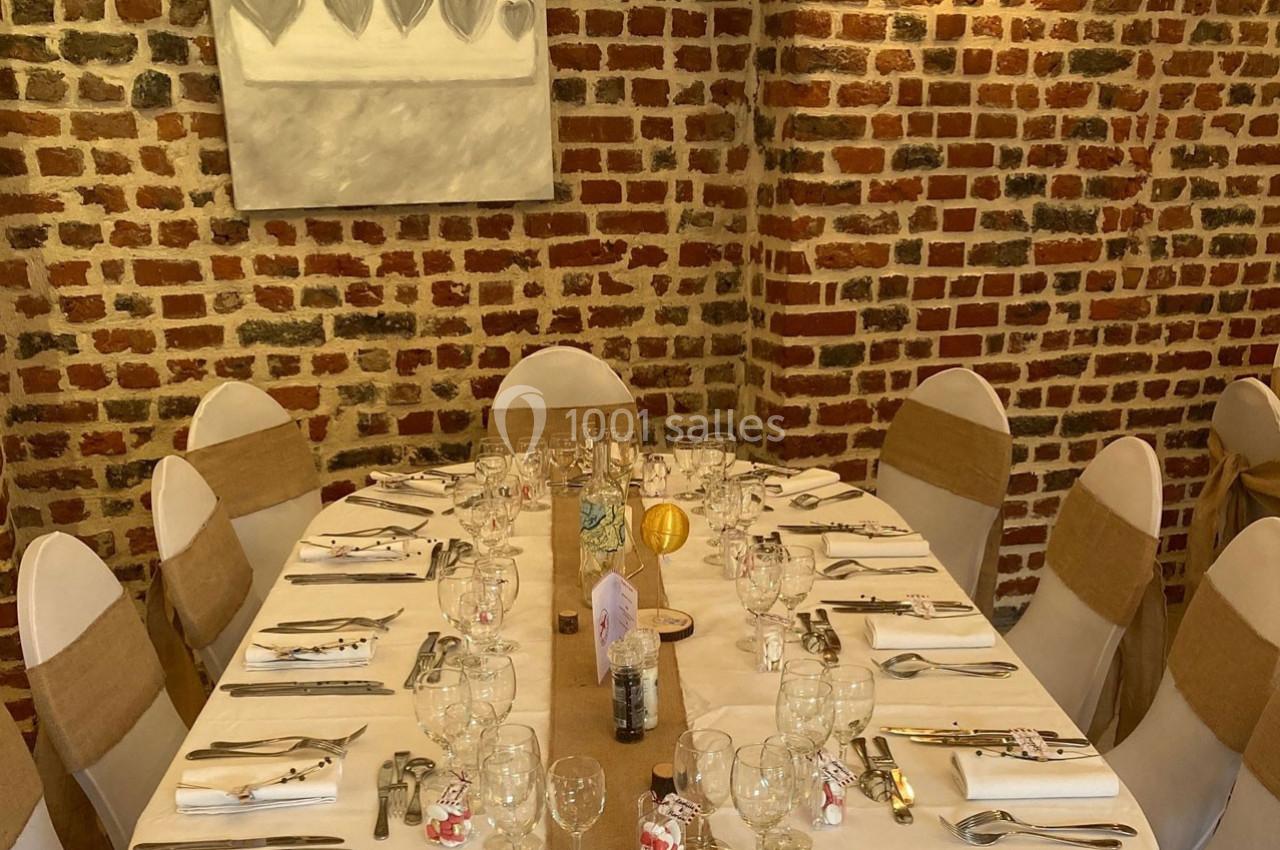 Table élégamment dressée pour un repas, entourée de chaises blanches avec nœuds beiges, devant un mur en briques.