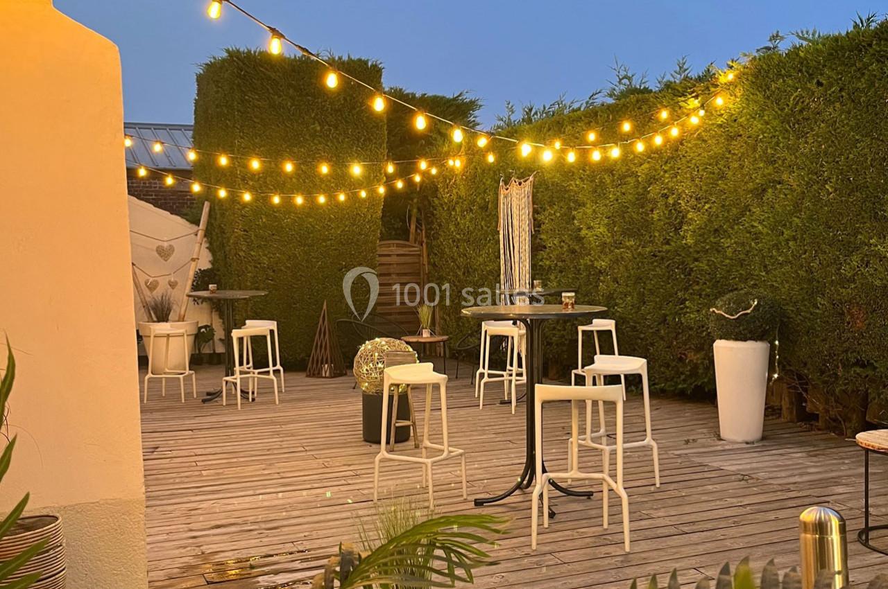 Terrasse en bois éclairée par des guirlandes lumineuses, avec tables hautes, tabourets et haies en arrière-plan.
