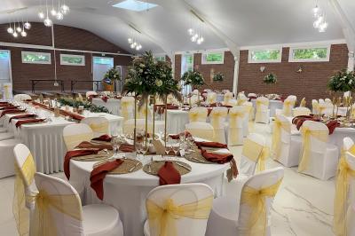 Table en bois décorée de bougies, fleurs et feuillage, entourée de chaises, sous une arche végétale dans une salle lumineuse.