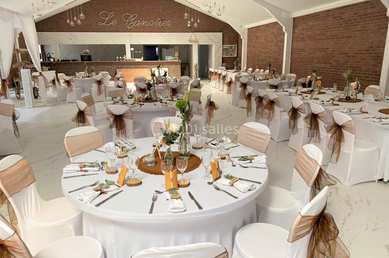 Salle de réception décorée avec des tables rondes, nappes blanches, chaises ornées de rubans et centre de table floral.