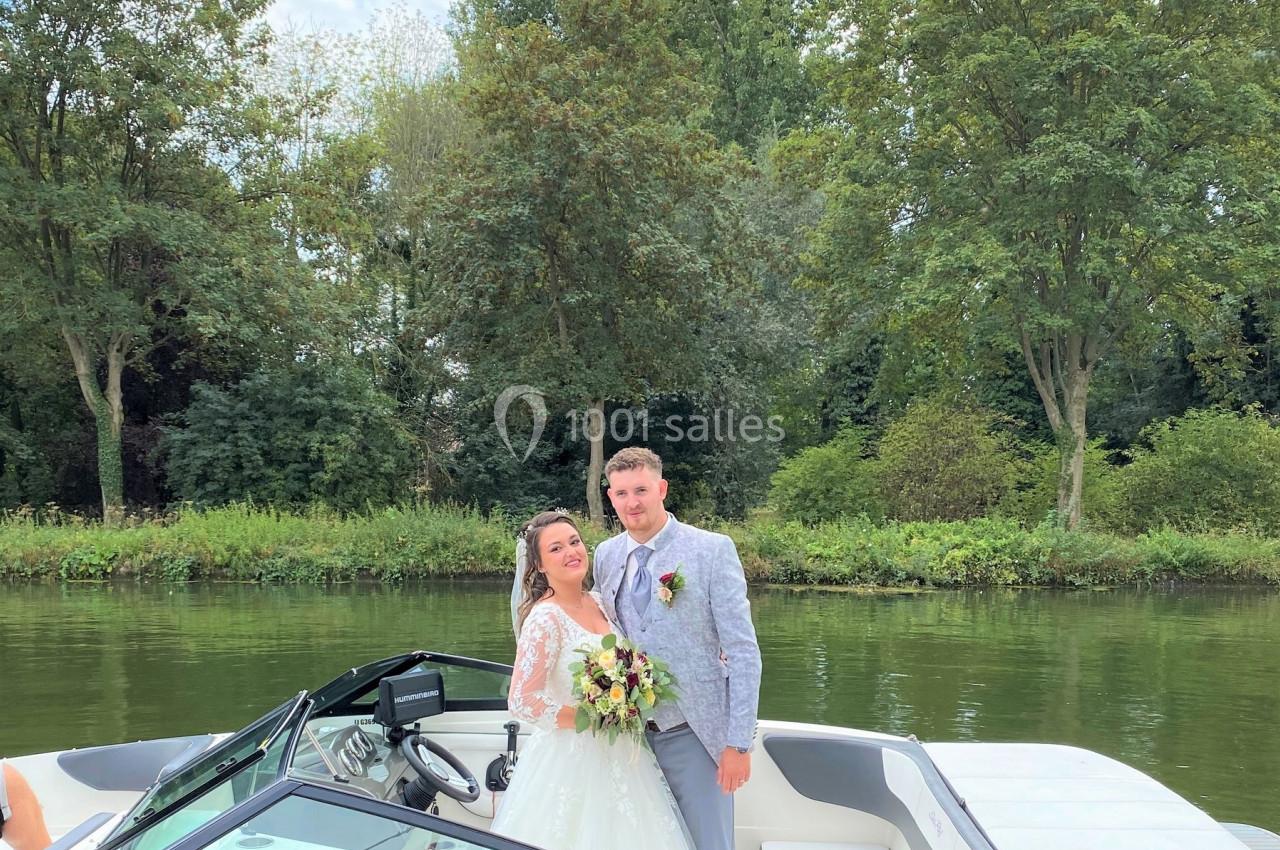 Un couple en tenue de mariage pose sur un bateau au bord d'une rivière entourée d'arbres.