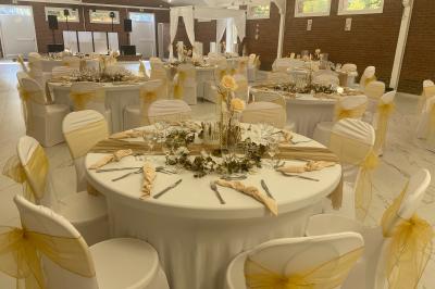 Table en bois décorée de bougies, fleurs et feuillage, entourée de chaises, sous une arche végétale dans une salle lumineuse.