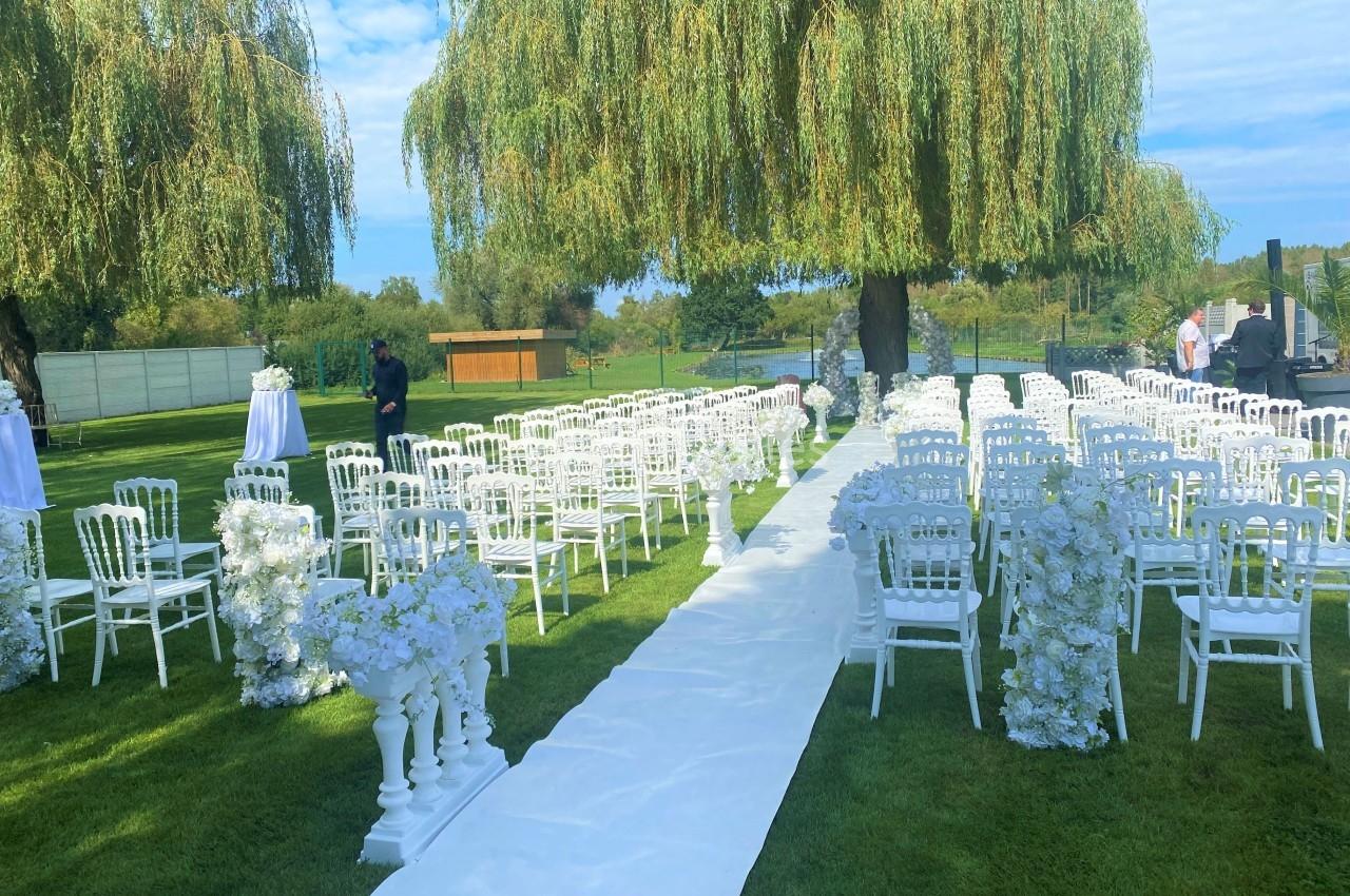 Allée centrale décorée de fleurs blanches entourée de chaises blanches, installée en extérieur sous des arbres.