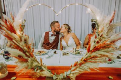 Table en bois décorée de bougies, fleurs et feuillage, entourée de chaises, sous une arche végétale dans une salle lumineuse.