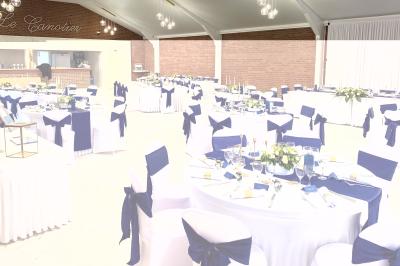 Table en bois décorée de bougies, fleurs et feuillage, entourée de chaises, sous une arche végétale dans une salle lumineuse.