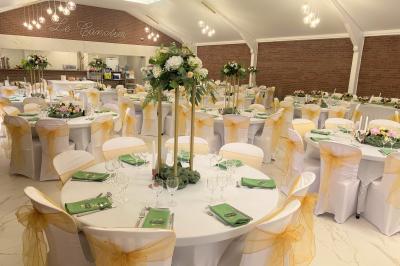 Table en bois décorée de bougies, fleurs et feuillage, entourée de chaises, sous une arche végétale dans une salle lumineuse.