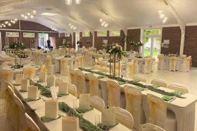 Table en bois décorée de bougies, fleurs et feuillage, entourée de chaises, sous une arche végétale dans une salle lumineuse.