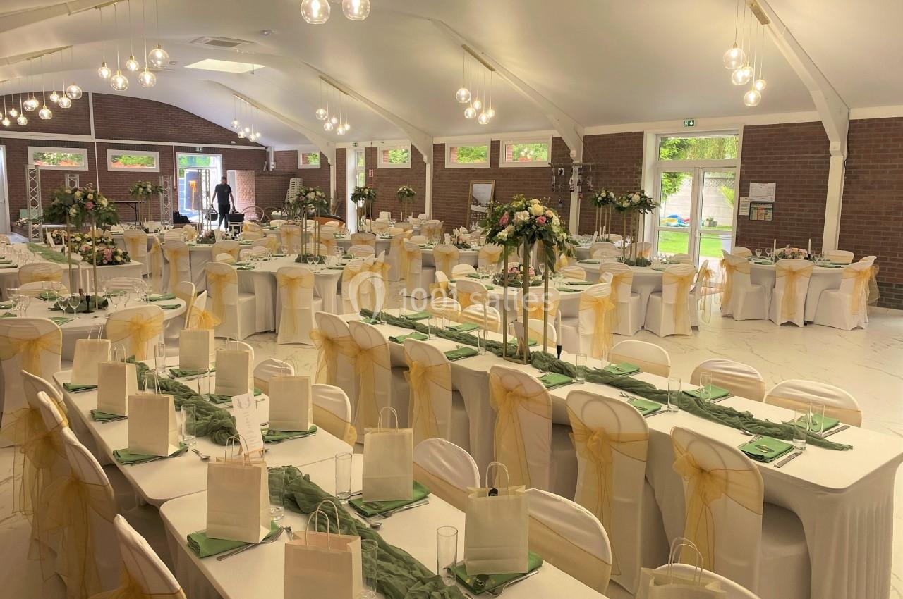 Salle de réception décorée pour un événement, avec des tables alignées, nappes blanches, chaises ornées de rubans dorés.