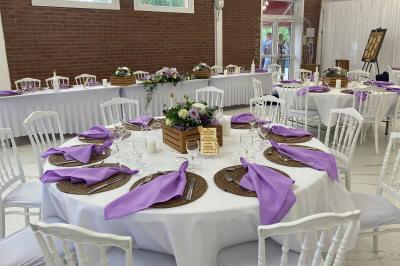 Table en bois décorée de bougies, fleurs et feuillage, entourée de chaises, sous une arche végétale dans une salle lumineuse.