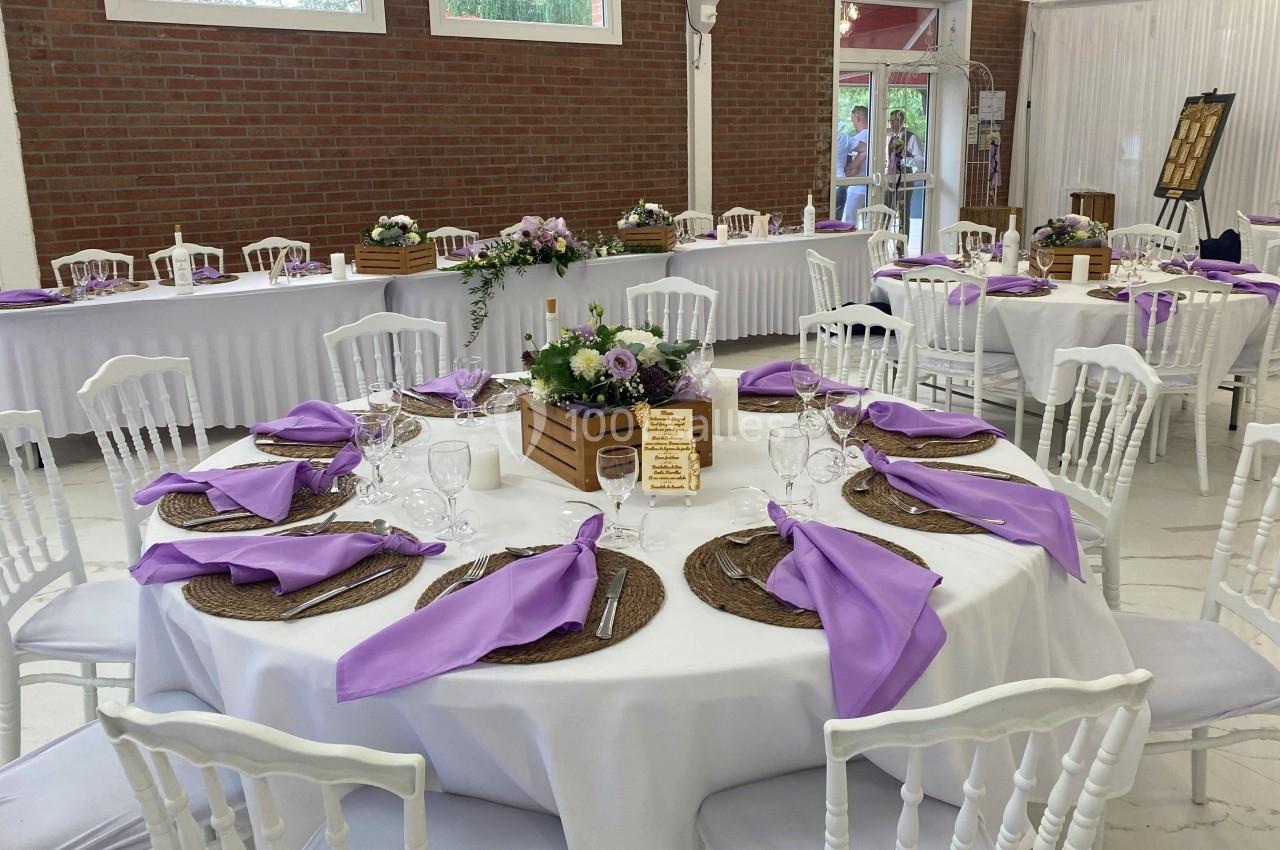 Table dressée pour un événement avec nappes blanches, serviettes violettes, décorations florales et chaises blanches.