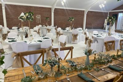 Table en bois décorée de bougies, fleurs et feuillage, entourée de chaises, sous une arche végétale dans une salle lumineuse.