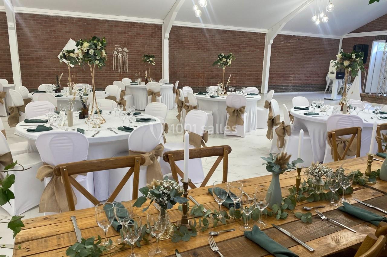 Salle de réception décorée pour un mariage, avec tables dressées, nappes blanches, chaises ornées de rubans et centres de…