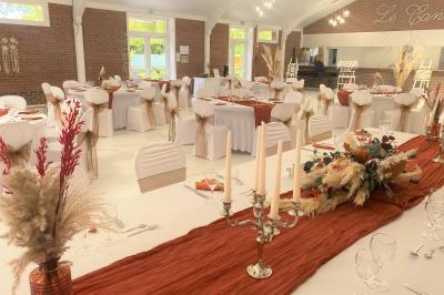 Table en bois décorée de bougies, fleurs et feuillage, entourée de chaises, sous une arche végétale dans une salle lumineuse.