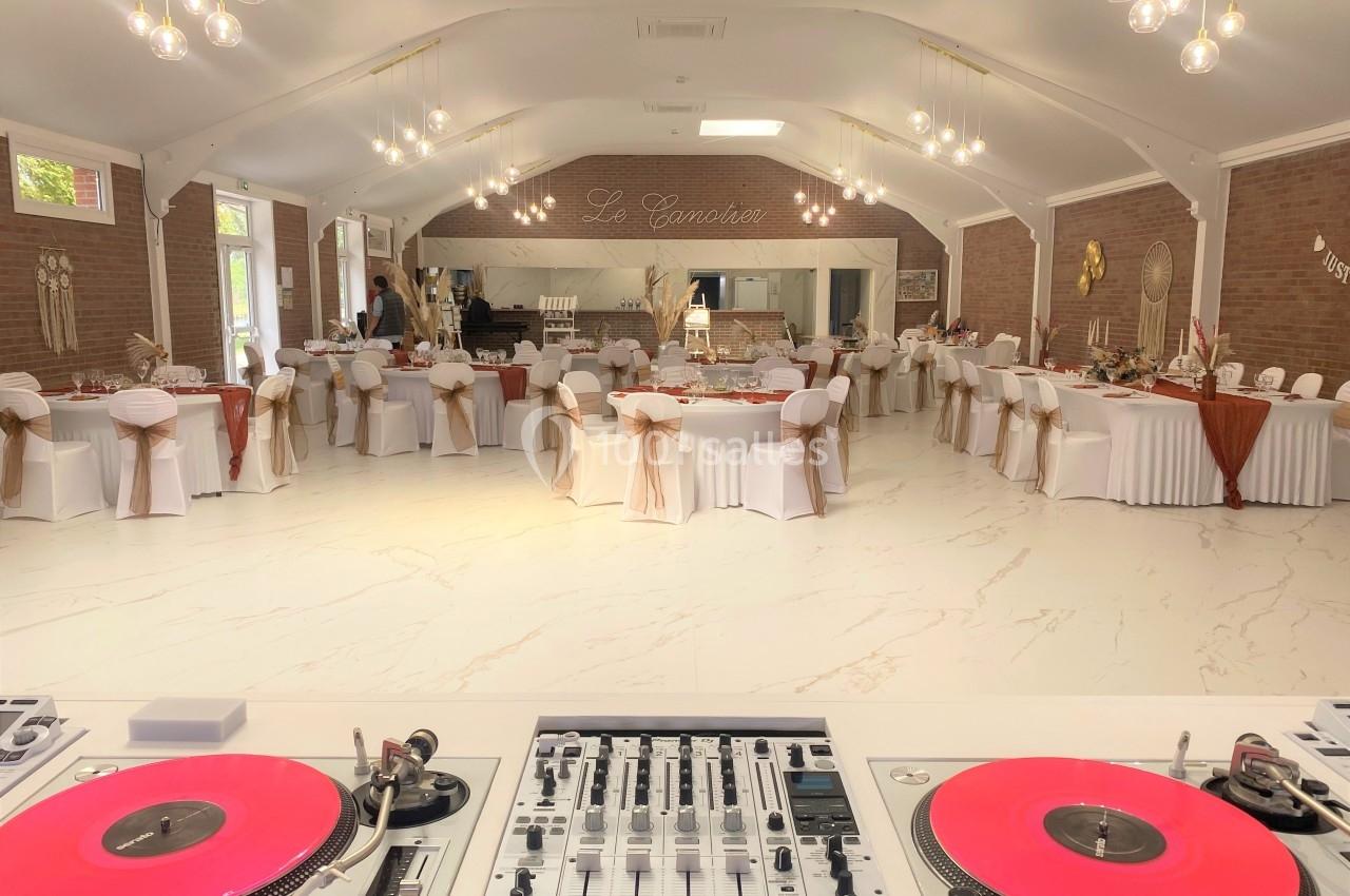 Salle de réception décorée avec des tables rondes et chaises blanches, vue depuis une table de mixage avec platines roses.