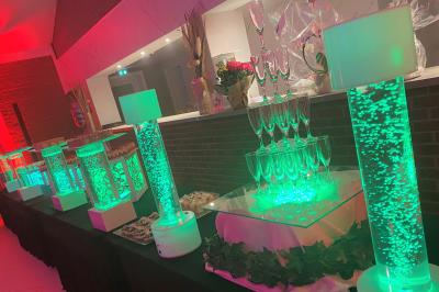 Table en bois décorée de bougies, fleurs et feuillage, entourée de chaises, sous une arche végétale dans une salle lumineuse.