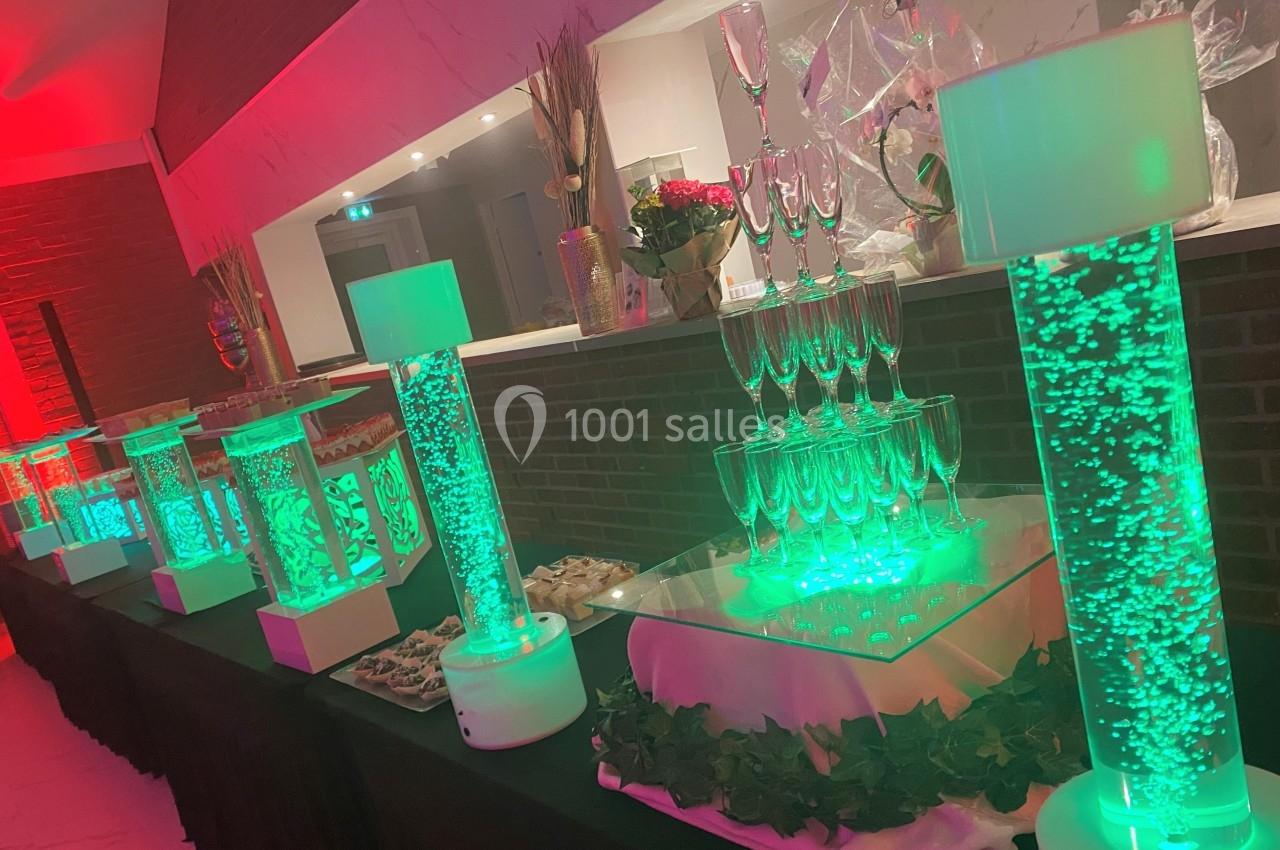 Table décorative avec colonnes lumineuses vertes, verres empilés et plateaux de nourriture dans un cadre festif.