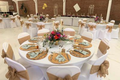 Table en bois décorée de bougies, fleurs et feuillage, entourée de chaises, sous une arche végétale dans une salle lumineuse.