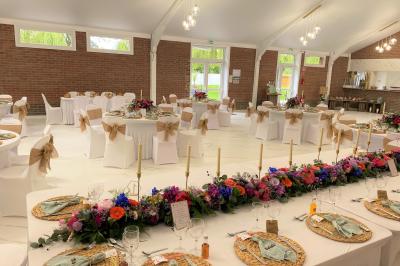 Table en bois décorée de bougies, fleurs et feuillage, entourée de chaises, sous une arche végétale dans une salle lumineuse.