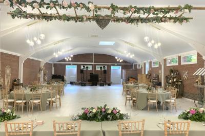 Table en bois décorée de bougies, fleurs et feuillage, entourée de chaises, sous une arche végétale dans une salle lumineuse.