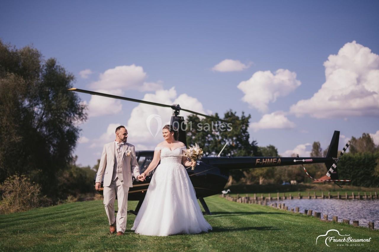 Un couple en tenue de mariage marche main dans la main près d'un hélicoptère, dans un cadre verdoyant et ensoleillé.