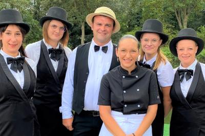 Table en bois décorée de bougies, fleurs et feuillage, entourée de chaises, sous une arche végétale dans une salle lumineuse.