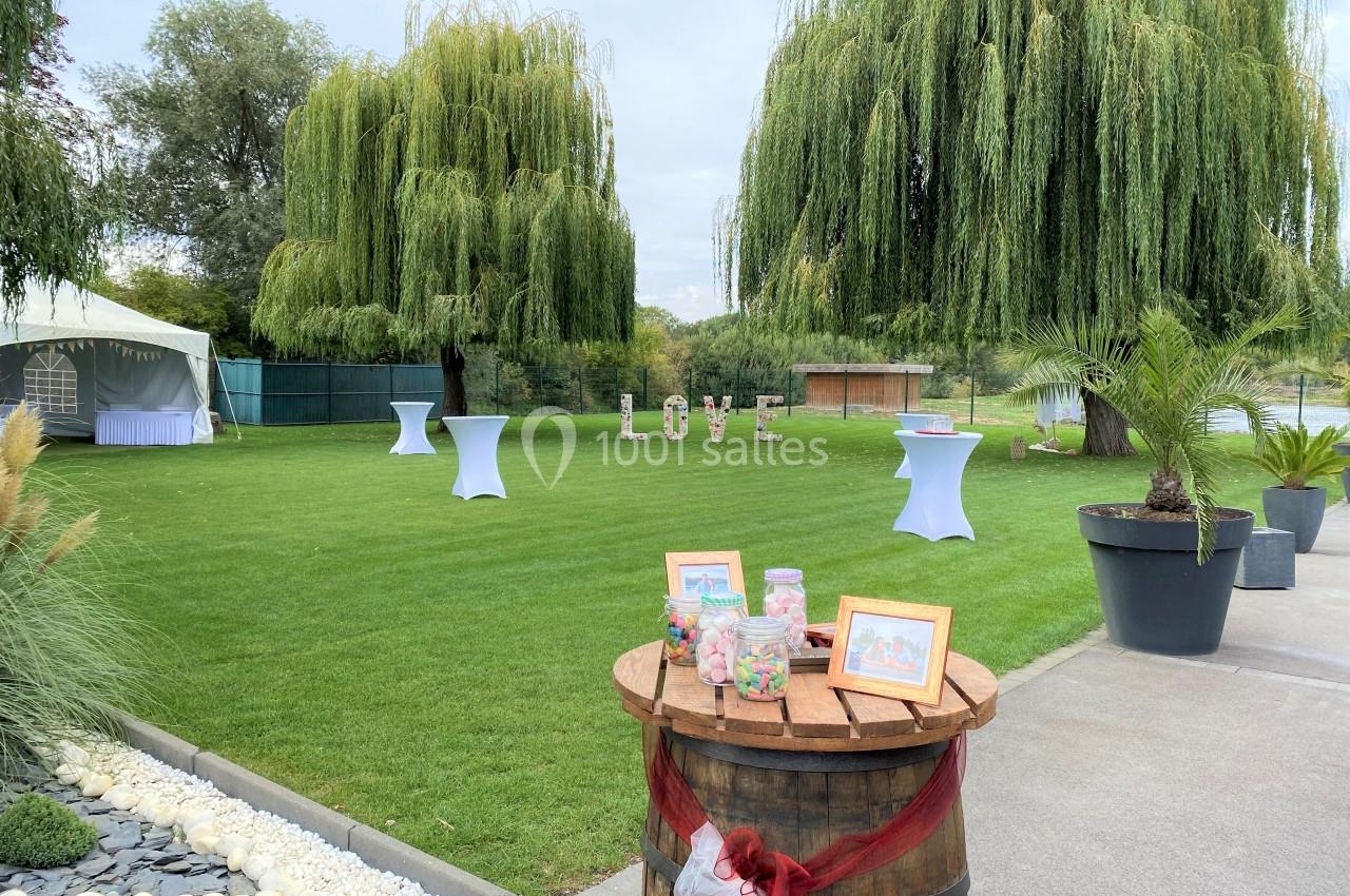 Jardin avec pelouse, tables hautes, décorations de mariage et grands saules pleureurs en arrière-plan.