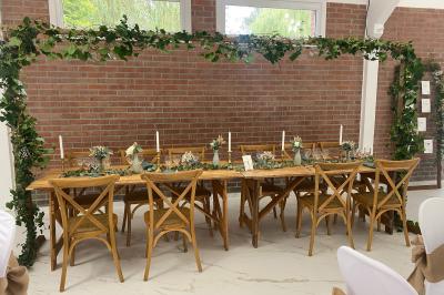 Table en bois décorée de bougies, fleurs et feuillage, entourée de chaises, sous une arche végétale dans une salle lumineuse.