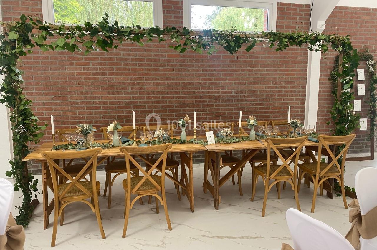 Table en bois décorée de bougies, fleurs et feuillage, entourée de chaises, sous une arche végétale dans une salle lumineuse.