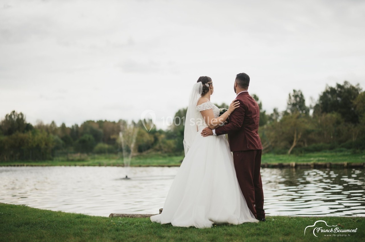 Un couple en tenue de mariage se tient près d'un étang, entouré de verdure et d'arbres sous un ciel nuageux.