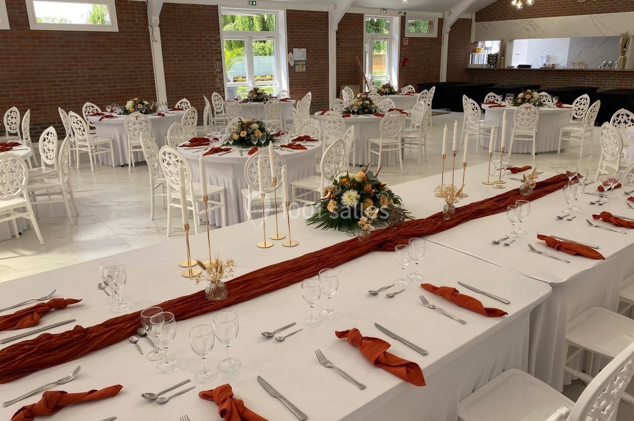 Salle de réception décorée avec des tables dressées, nappes blanches, serviettes orange et centres de table floraux.