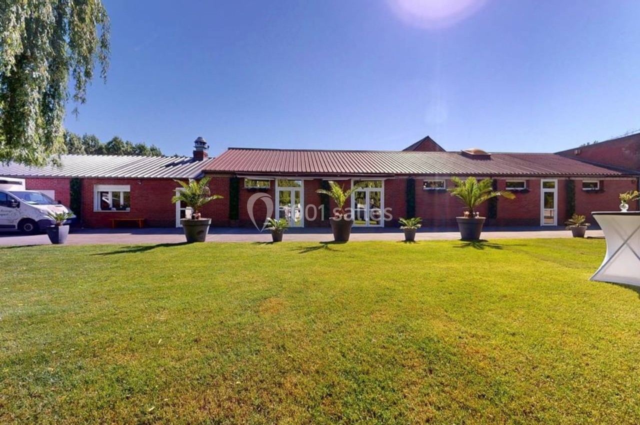 Bâtiment en briques rouges avec toit en tôle, entouré de pelouse et de plantes en pots sous un ciel dégagé.