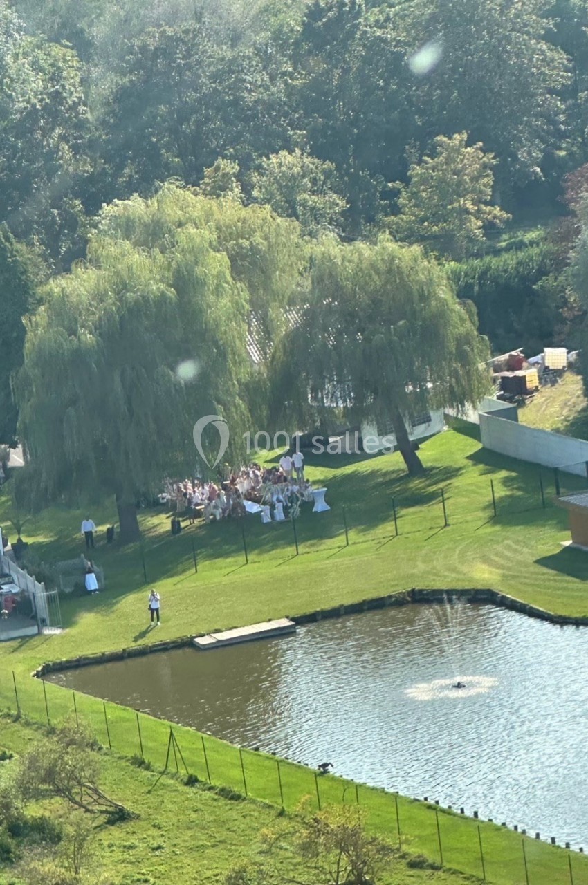 Vue aérienne d'un rassemblement en plein air sous des arbres près d'un étang avec une fontaine, dans un cadre verdoyant.