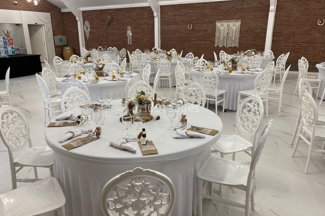 Salle de réception décorée avec des tables rondes dressées, chaises blanches et éléments de décoration bohème.