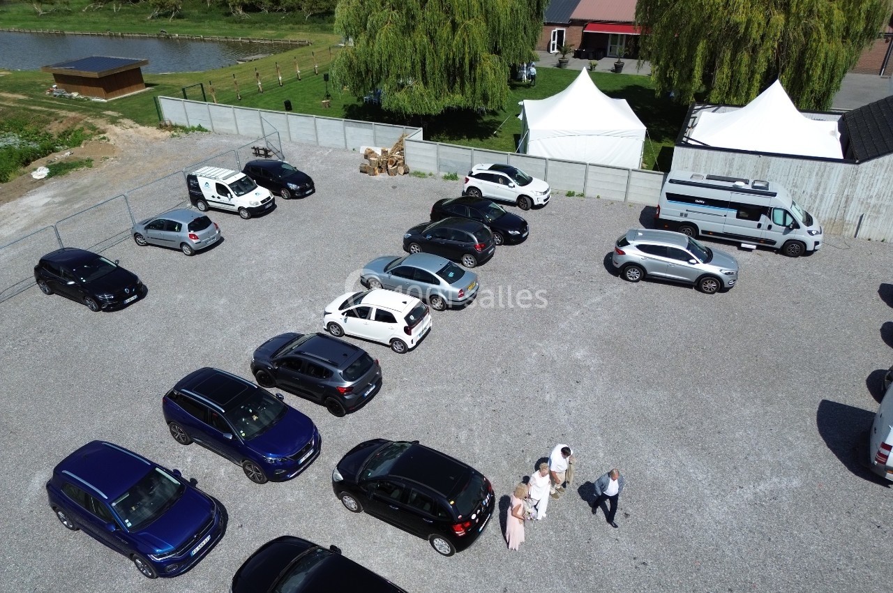 Vue aérienne d'un parking en gravier avec plusieurs voitures stationnées, des tentes blanches et un bâtiment en arrière-plan.