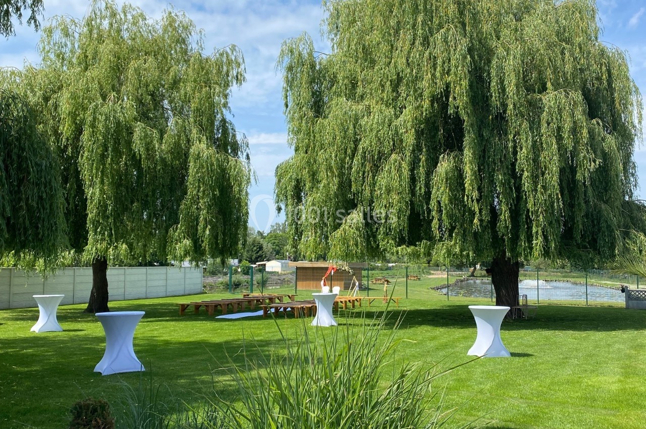 Jardin verdoyant avec deux grands saules, tables hautes blanches et bancs en bois, sous un ciel dégagé.