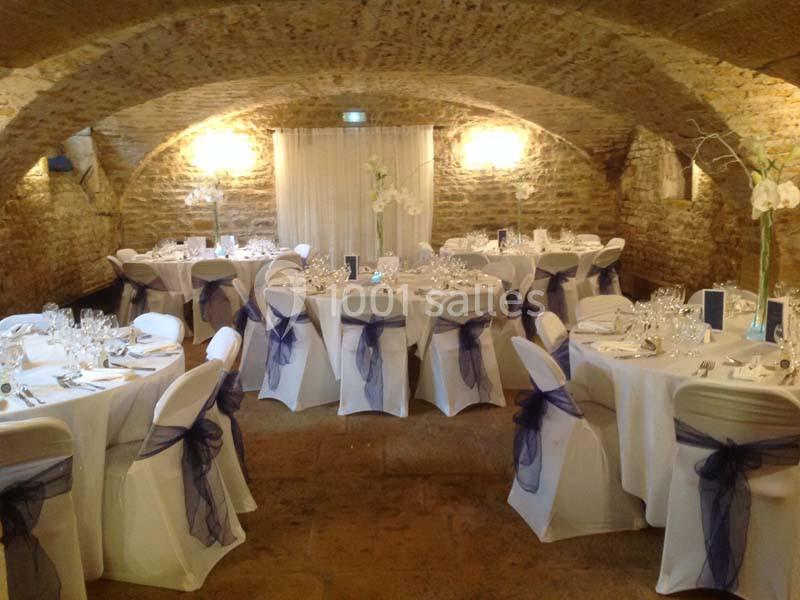Salle voûtée en pierre aménagée pour un événement, avec tables rondes dressées et chaises décorées de nœuds bleus.