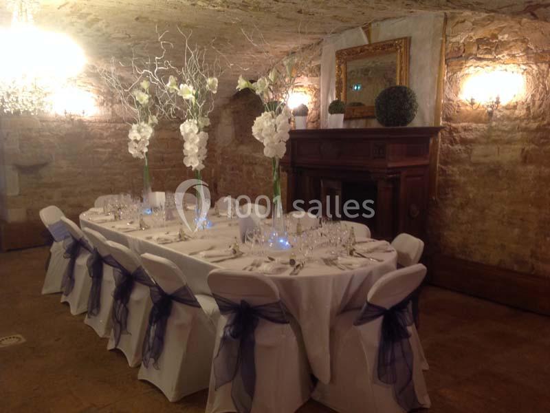 Table élégamment dressée dans une salle voûtée en pierre, décorée de fleurs blanches et de chaises ornées de rubans.